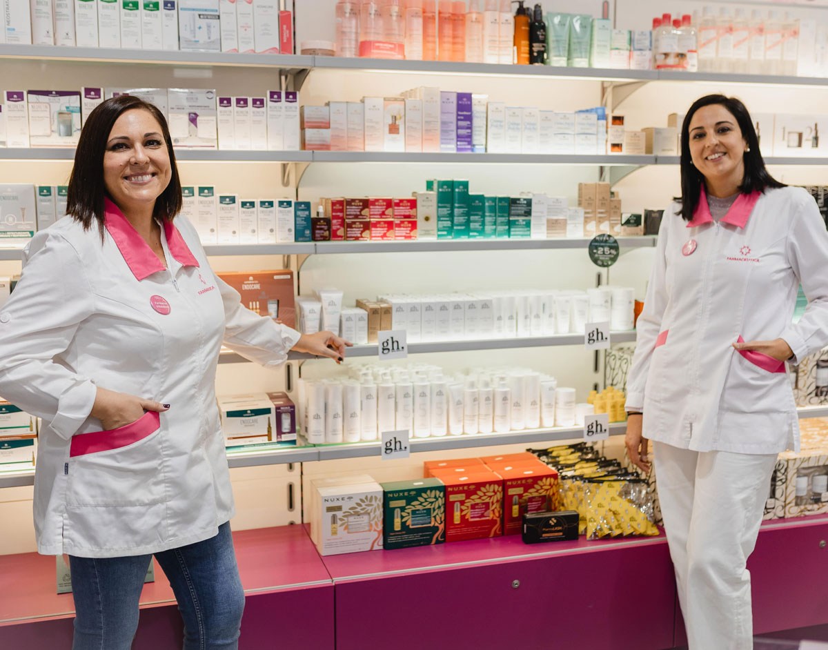 Se ve una imagen con dos farmacéuticas apoyadas en una estanteria de su farmacia